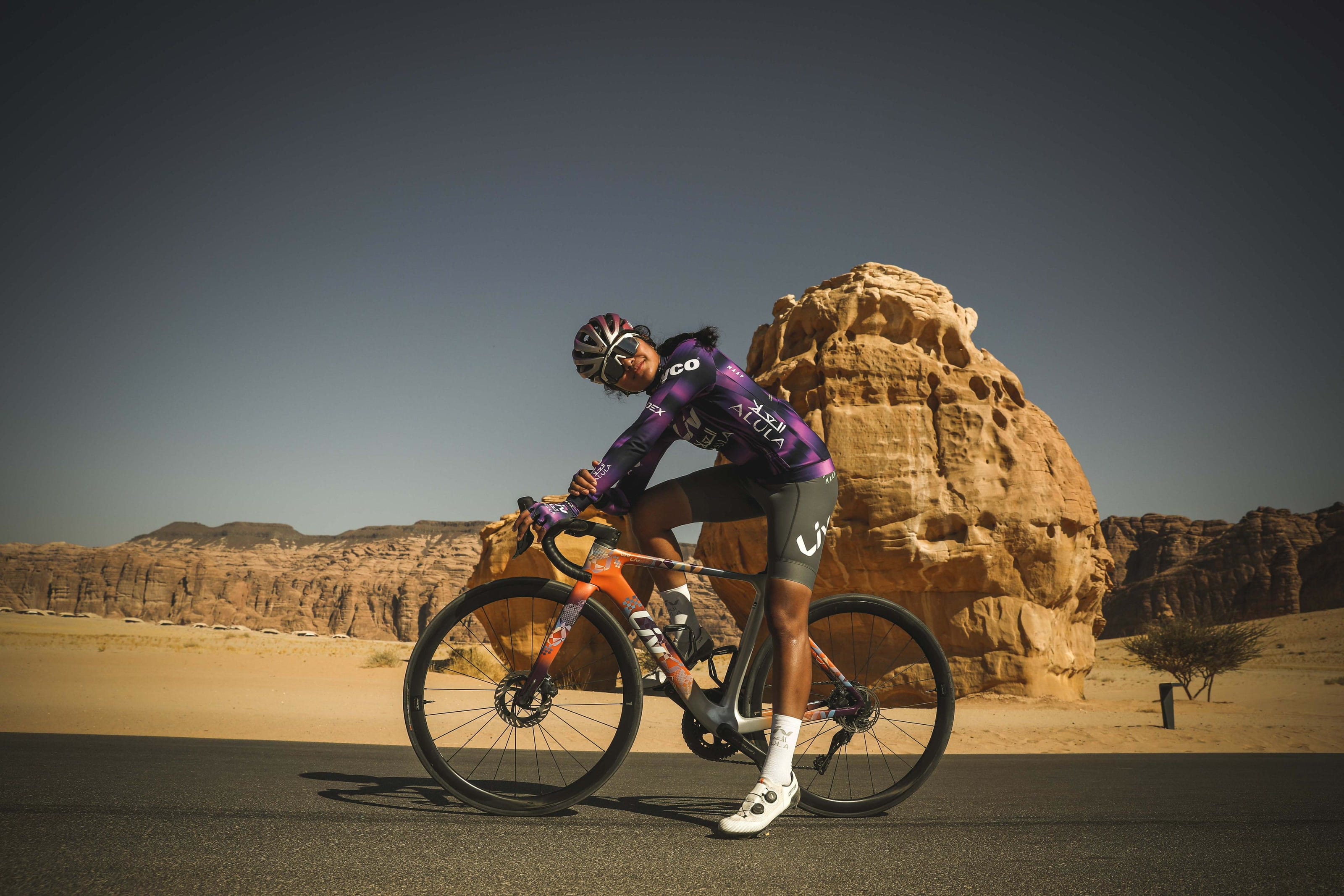 Moroj Adil in profile with her bike on a road in AlUla, sandstone behind her